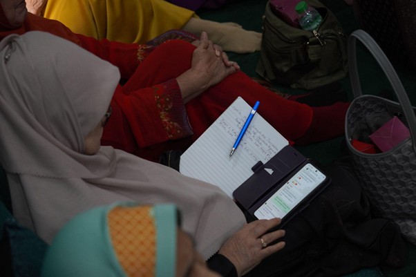 Woman in a beige hijab writes notes in a notebook during a gathering, with a blue pen nearby and a phone in hand nearby pieces of colorful clothing in the background.