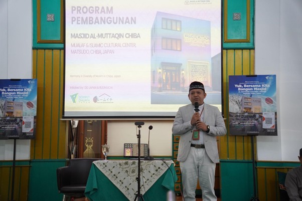 Speaker in a light suit with a microphone presenting at a mosque construction program, projection screen behind him shows related title text on Indonesian slide.