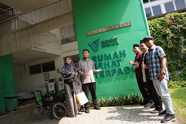 senseisugimotokunjungismart12 dompet dhuafa Five adults pose outside a green health center with a sign that reads 'Rumah Sehat Terpadu' and wheelchairs nearby.