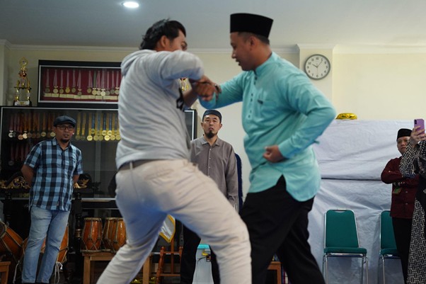 senseisugimotokunjungismart7 dompet dhuafa Two men sparring in a room as onlookers watch, with trophies displayed in the background and a clock on the wall.