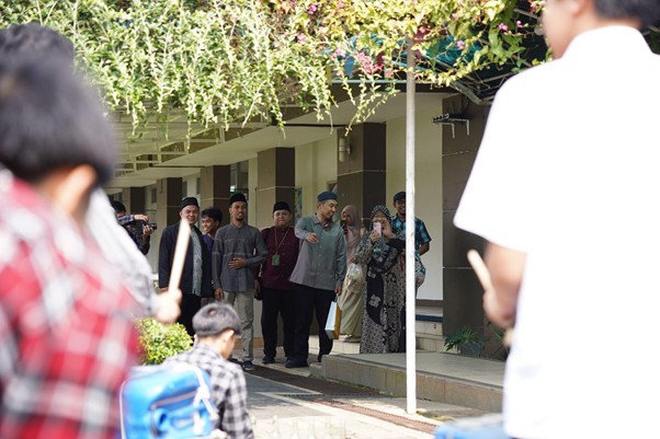 senseikesmart2 dompet dhuafa People greet each other at the entrance of a building, with onlookers and a few waving hello; a student with a blue backpack sits in the foreground.