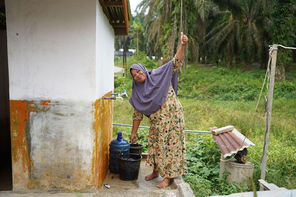 sumur bor di aceh tamiang 1 dompet dhuafa Barefoot woman in a purple headscarf and floral dress stands on a concrete platform and uses a rope to lift a bucket at an outdoor water tap, with a white-and-orange building beside her and lush fields in the background.