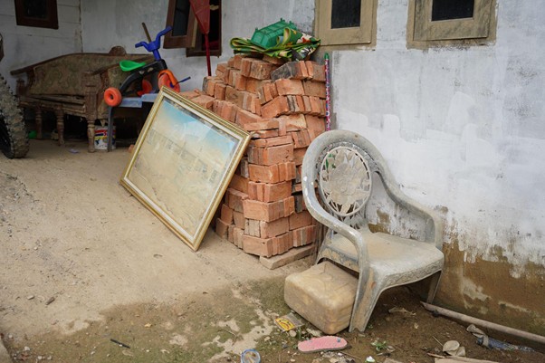 sumur bor di aceh tamiang 2 dompet dhuafa Outdoor corner with a stack of red bricks, a framed painting leaning against them, a gray plastic chair, and scattered debris