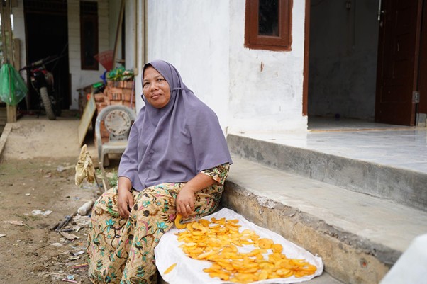sumur bor di aceh tamiang 4 dompet dhuafa Woman wearing a purple hijab sits on a doorstep with a tray of sun-drying orange mango slices beside her, in a rural yard.