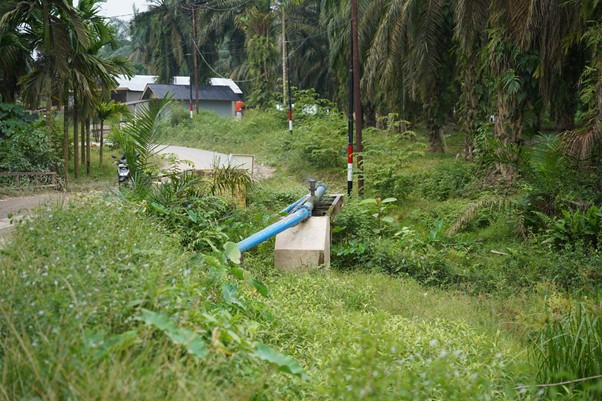 sumur bor di aceh tamiang 5 dompet dhuafa Blue irrigation pipe resting on concrete supports in a lush rural area with palm trees and a small building in the background.