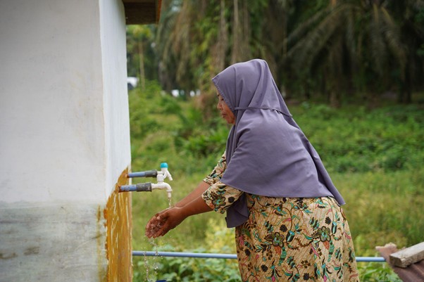 sumur bor di aceh tamiang 7 dompet dhuafa Woman in a purple headscarf washes her hands at an outdoor faucet mounted on a white wall in a green, rural setting.