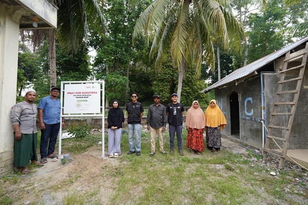 sumur bor di bireun aceh 1 dompet dhuafa Nine people stand outdoors beside a sign that reads 'Pray for Sumatera' in a tropical yard with palm trees and simple buildings.
