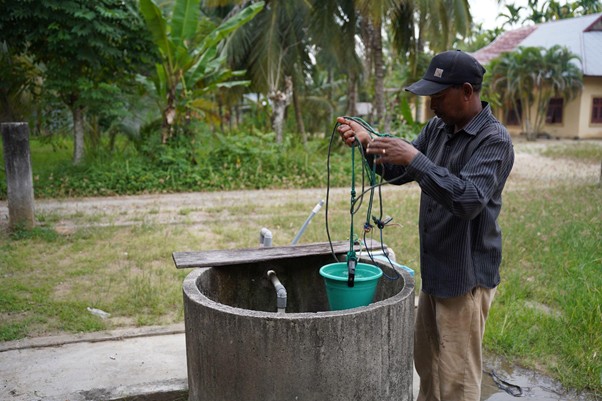 sumur bor di bireun aceh 2 dompet dhuafa Man wearing a cap untangles a rope, lowering a green bucket into a concrete well in a rural backyard.
