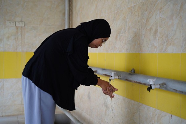 sumur bor di bireun aceh 6 dompet dhuafa Woman wearing a black hijab washing her hands at a wall-mounted faucet in a tiled room, water running from the tap.