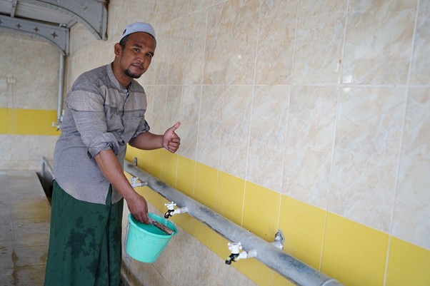 sumur bor di bireun aceh 8 dompet dhuafa Man in a cap stands by tiled wall, holding a green bucket under outdoor taps and giving a thumbs up while washing.