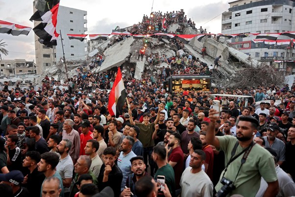 Large crowd packs a rubble-strewn street, waving red-white-black flags amidst collapsed buildings and dusted skyline.