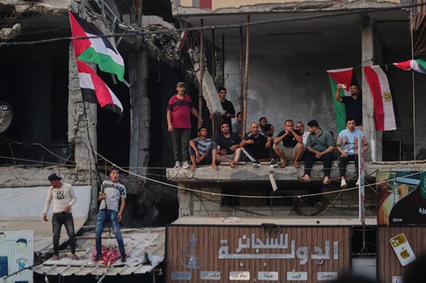 tokoh kemanusiaan gaza 3 dompet dhuafa Group of men sitting and standing on the edge of a damaged concrete building, Palestinian flags hanging from the upper level above a rubble-filled street.