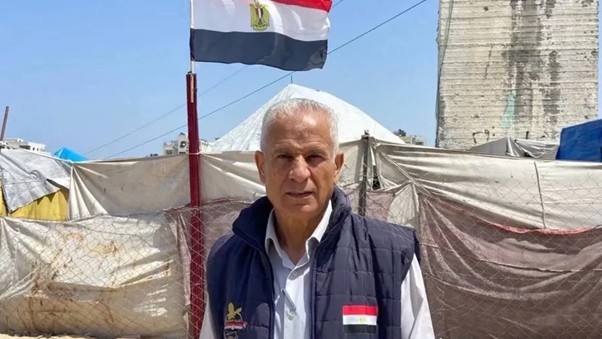 tokoh kemanusiaan gaza 4 dompet dhuafa Middle-aged man in a dark vest over a white shirt stands in a temporary camp with tents; an Egyptian flag flies overhead.