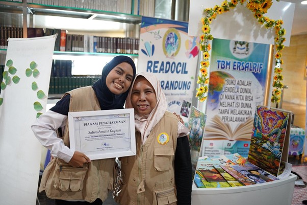 Two women in beige vests smile at a booth, one holding a certificate, with literacy banners in the background.
