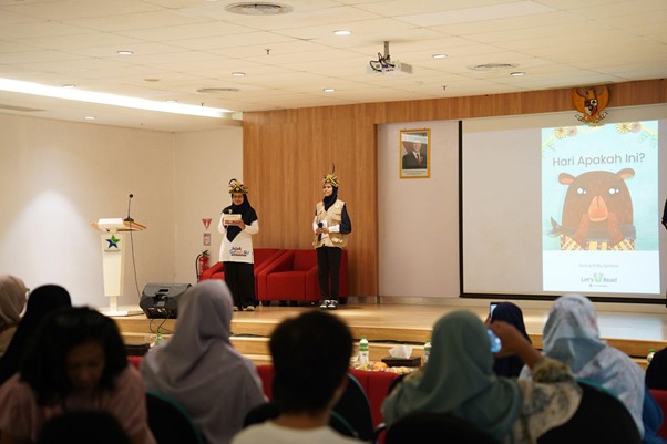 yatim penakluk speech delay 2 dompet dhuafa Two presenters in animal-themed costumes stand on a stage addressing an audience in a conference room, with a cartoon bear slide projected on the screen to the right.