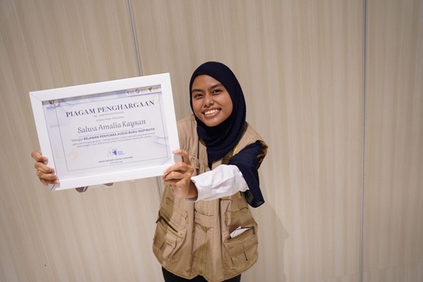 yatim penakluk speech delay 5 dompet dhuafa Smiling woman wearing a hijab and beige utility vest holds up a framed award certificate toward the camera.