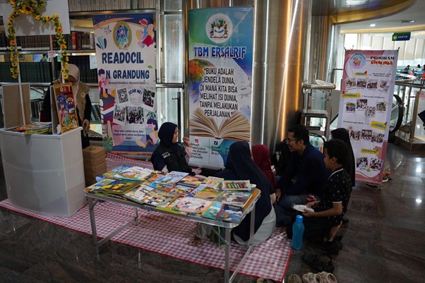 Group of people sit on a checkered mat around a low table full of books at a reading event, banners behind them with Indonesian text.