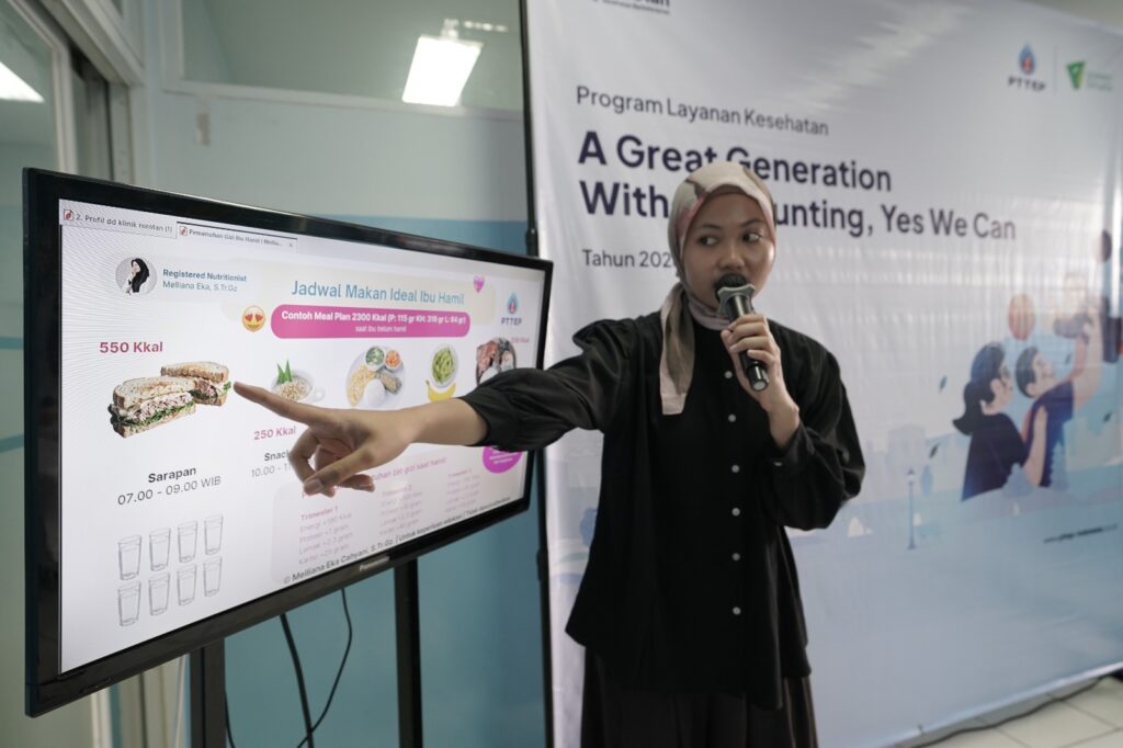 yoga ibu hamil 1 dompet dhuafa Presenter with a microphone points at a large screen displaying a meal-plan infographic for a health program.