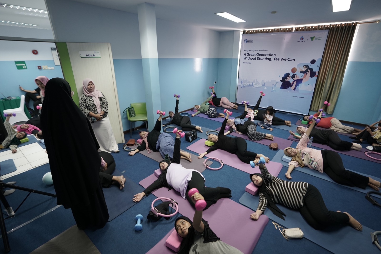 yoga ibu hamil 2 dompet dhuafa Women in a group fitness class lying on mats and lifting pink dumbbells in a room with a health banner in the background.
